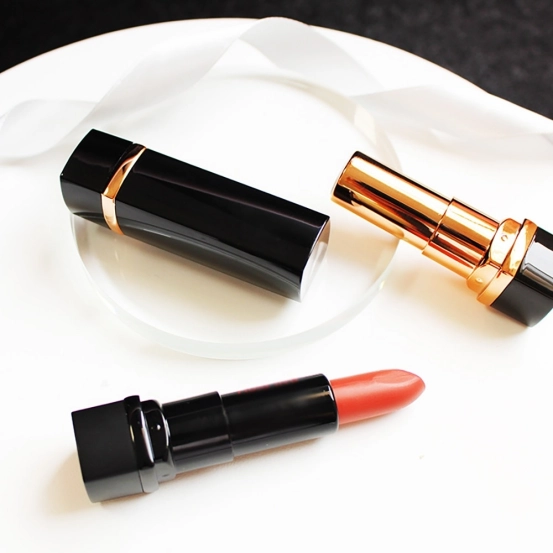 Glossy black lipstick tubes with gold accents shown closed, open, and extended with a coral lipstick bullet on a white display surface.